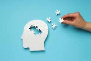 Puzzle eines menschlichen Kopfs im Profil, vier Puzzleteile sind nicht eingesetzt, drei liegen an der Seite, eines in einer Hand. Blauer Hintergrund.
