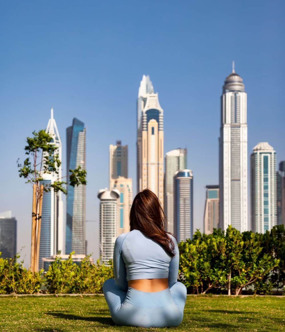 Brünette Frau in blauer Sportkleidung hockt von Hinten im Gras mit Blick auf Bäume und die Dubai-Skyline unter blauem Himmel.