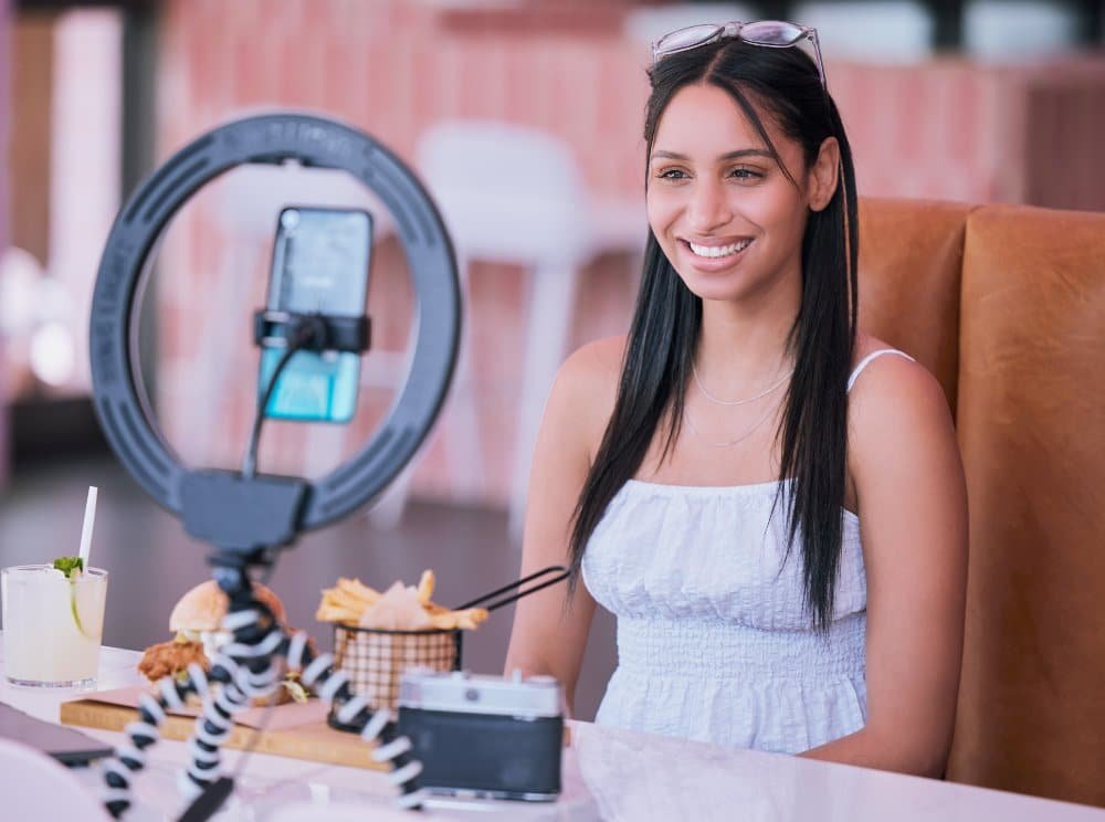 Lächelnde, dunkelhaarige Frau mit Sonnenbrille auf Kopf und weißem Topf, sitzt in einem Restaurant mit Burger, Pommes und Limo am Tisch und filmt sich mit Handy und Ringlicht. Rötlich-braune Wand und weiße Hocker im Hintergrund.