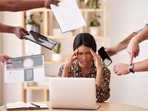 Schwarzhaarige Frau mit Zopf in braun-beige gemusterter Bluse sitzt mit Kopf auf Händen gestützt vor Computer. Um sie herum 5 Handpaare mit Armbanduhr, Handy, Tablet und zwei Notizblöcken. Helle Wand mit Regal verschwommen im Hintergrund.