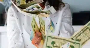 Frau vom Hals bis zum Brustbereich in weißem Pulli mit schwarzen Punkten wirft Euro- und Dollarscheine in die Luft. Verschwommener Hintergrund mit weißer Wand und schwarzen Sesseln.