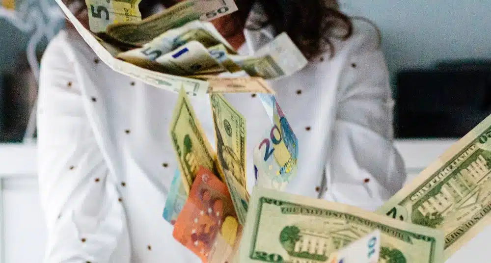 Frau vom Hals bis zum Brustbereich in weißem Pulli mit schwarzen Punkten wirft Euro- und Dollarscheine in die Luft. Verschwommener Hintergrund mit weißer Wand und schwarzen Sesseln.