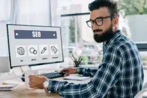Dunkelhaariger Mann mit Drei-Tage-Bart, Brille und blau kariertem Hemd sitzt am Computerbildschirm, auf dem SEO steht und blickt über die Schulter. Fenster im Hintergrund.