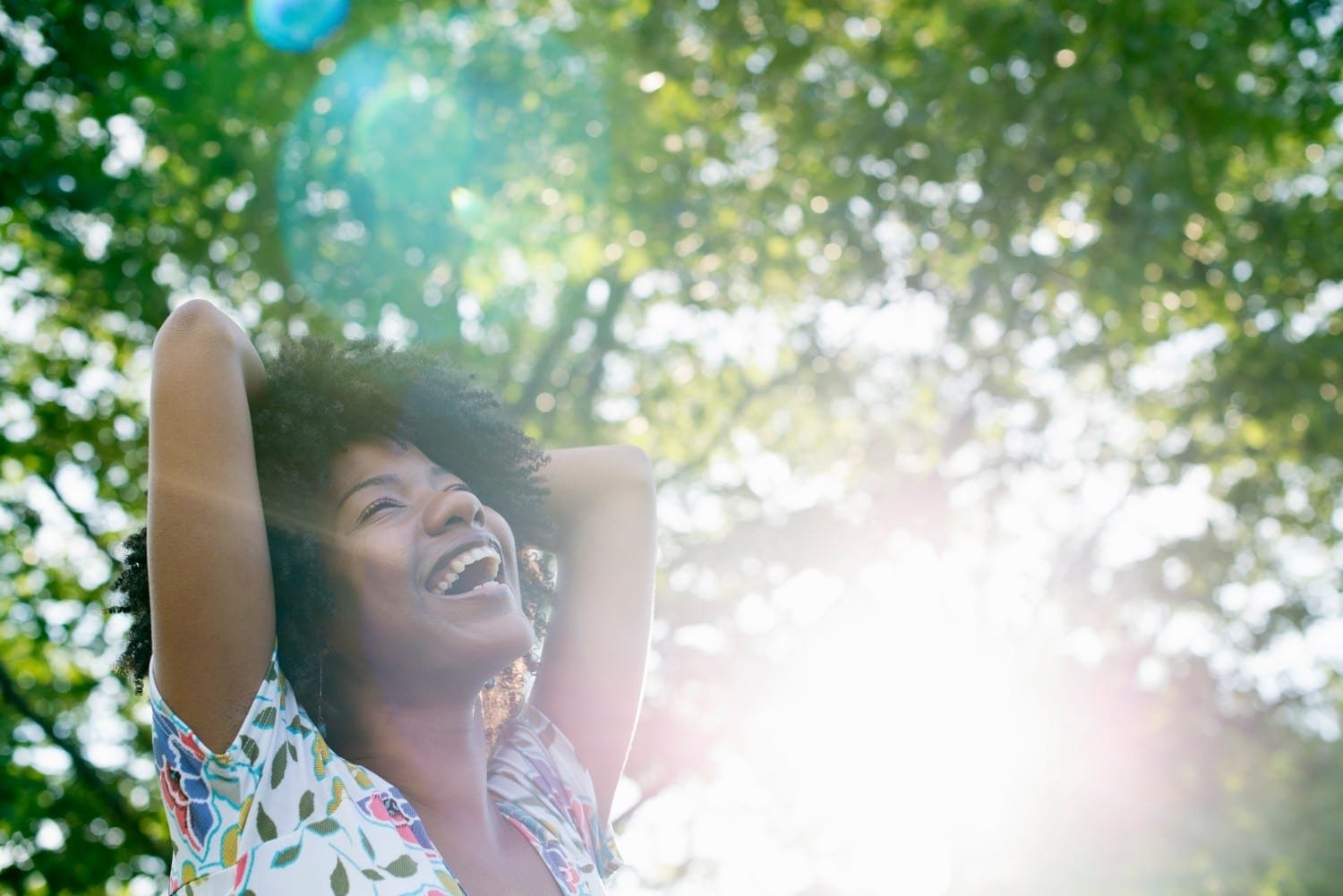 Lachende, schwarzhaarige Frau mit geblümten T-Shirt hält Arme auf den Kopf und blickt lachend auf das Blattwerk über ihr.
