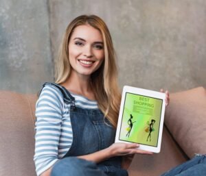 Blonde Frau mit blauer Jeans-Latzhose, gestreiften Shirts und Chucks sitzt im Schneidersitz auf braunen Kissen und zeigt lächelnd ein E-Book mit grünem Cover. Graue Wand im Hintergrund.