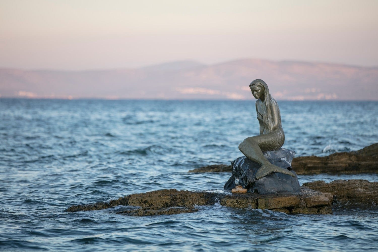 Bronzestatue “Die kleine Meerjungfrau” auf Fels am Meer. Hügel am Horizont.