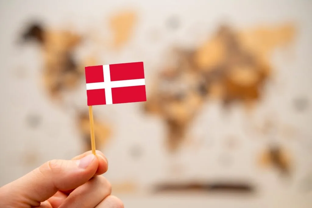 Hand, die Dänemark-Flagge auf Zahnstocher zwischen den Fingern hält. Goldene Weltkarte verschwommen im Hintergrund.