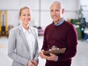Eine blonde Frau in grauem Business-Kostüm und ein grauhaariger Mann in rotem Pullover mit Kragen und Klemmbrett in der Hand stehen in einem hellen Gebäude und lächeln.