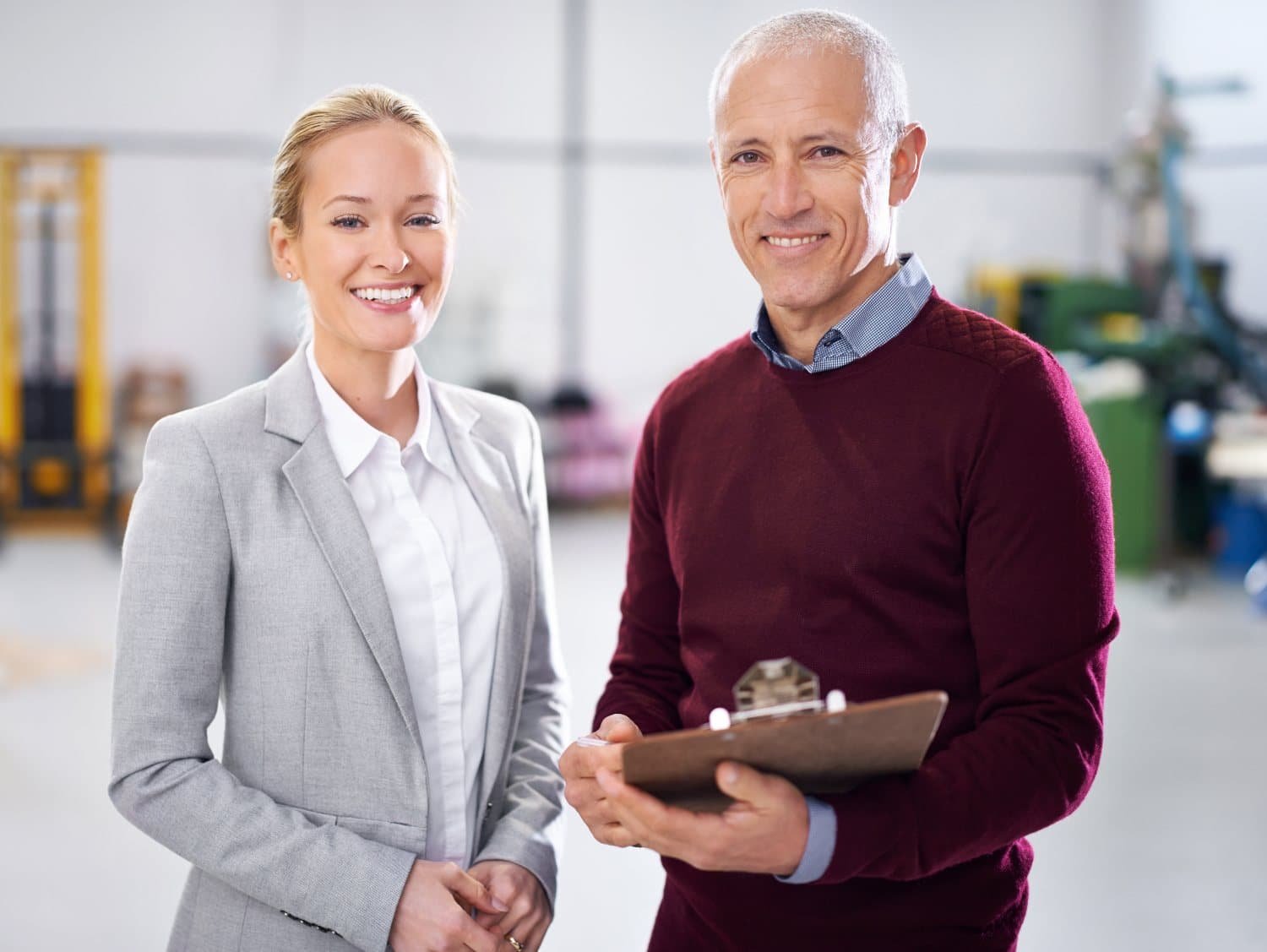 Eine blonde Frau in grauem Business-Kostüm und ein grauhaariger Mann in rotem Pullover mit Kragen und Klemmbrett in der Hand stehen in einem hellen Gebäude und lächeln.