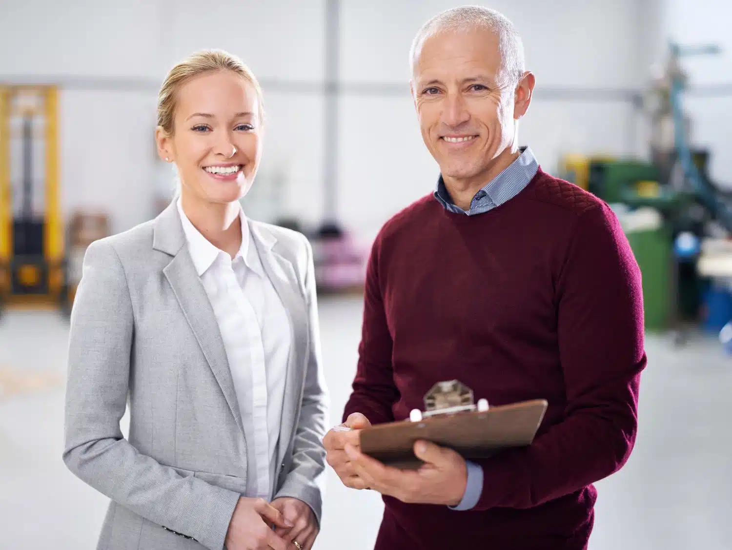 Eine blonde Frau in grauem Business-Kostüm und ein grauhaariger Mann in rotem Pullover mit Kragen und Klemmbrett in der Hand stehen in einem hellen Gebäude und lächeln.