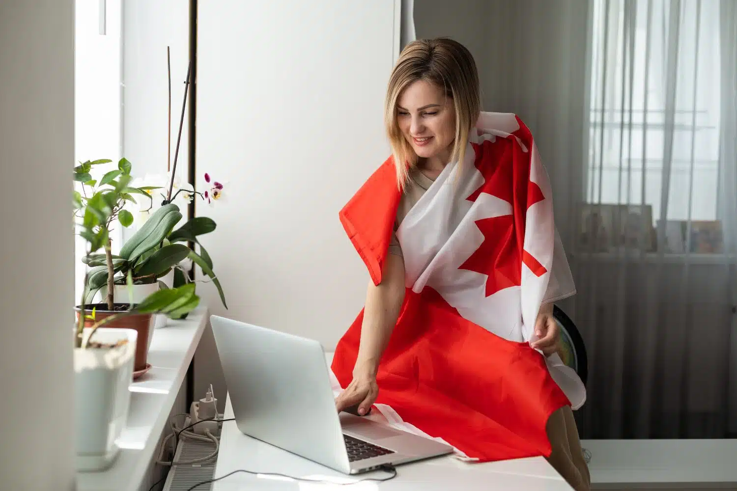 Blonde Frau, in Kanada-Flagge gehüllt, steht lächelnd am Laptop. Weißer Schreibtisch und Gardinen sowie Zimmerpflanzen im Hintergrund.