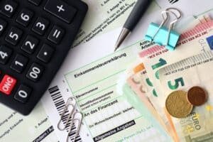 Einkommensteuererklärungsformular, darauf liegen zwei Büroklammern, eine blaue Heftklammer, ein Kugelschreiber, ein Taschenrechner und Euroscheine sowie zwei Münzen.