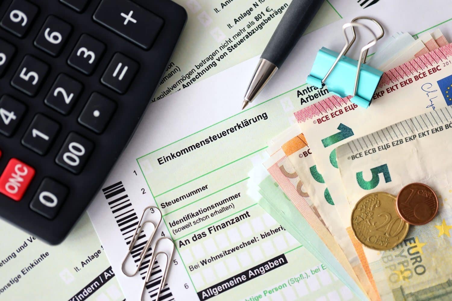 Einkommensteuererklärungsformular, darauf liegen zwei Büroklammern, eine blaue Heftklammer, ein Kugelschreiber, ein Taschenrechner und Euroscheine sowie zwei Münzen.