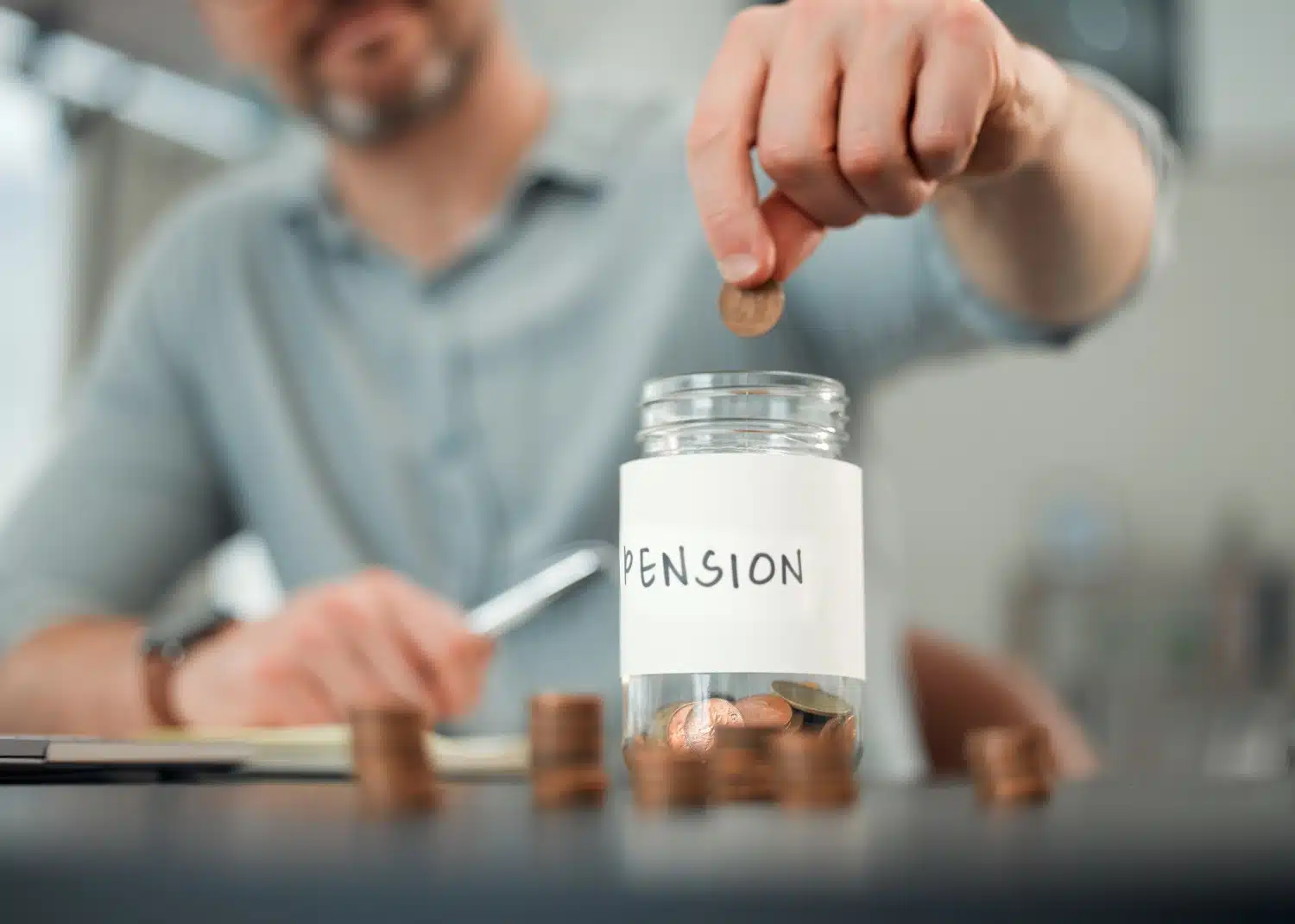 Verschwommener Mann vom Brustbereich bis zum Kinn, mit grauem Bart und blauem Hemd, sitzt am Schreibtisch und wirft Münzen in ein Glas, auf dem Pension steht. Stapel mit Kupfermünzen und Laptop daneben.
