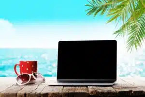 Laptop, rote Tasse mit weißen Punkten und rosa Sonnenbrille auf dunklem Holztisch. Blauer Himmel, Meer und Palme im Hintergrund.