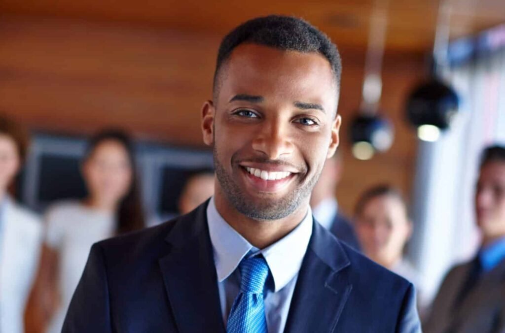 Schwarzhaariger Mann mit Drei-Tage-Bart in blauem Anzug lächelt in Kamera. Hinter ihm verschwommen sechs Personen, dahinter eine braune Wand ,darüber eine braune Zimmerdecke mit hängenden, schwarzen Deckenlampen.