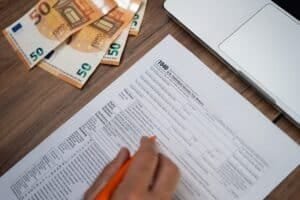 Eine männliche Hand mit Bleistift füllt ein Steuerformular am Schreibtisch vor Laptop aus, links daneben 50-Euro-Scheine gefächert.