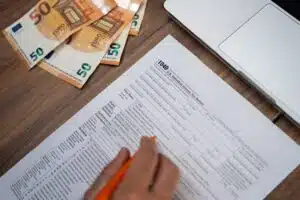 Eine männliche Hand mit Bleistift füllt ein Steuerformular am Schreibtisch vor Laptop aus, links daneben 50-Euro-Scheine gefächert.