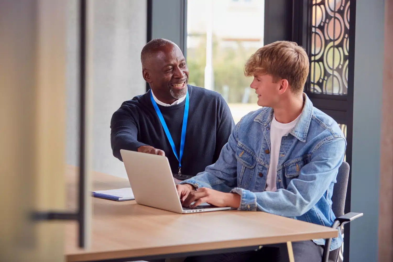 Älterer Mann in dunkelblauem Pullover und hellblauem Stoffanhänger um Hals, erklärt jüngeren blonden Mann in hellblauer Jeansjacke und weißem Shirt am Schreibtisch vor Laptop etwas. Fenster, brauner Rahmen mit Muster und weiße Wand im Hintergrund.