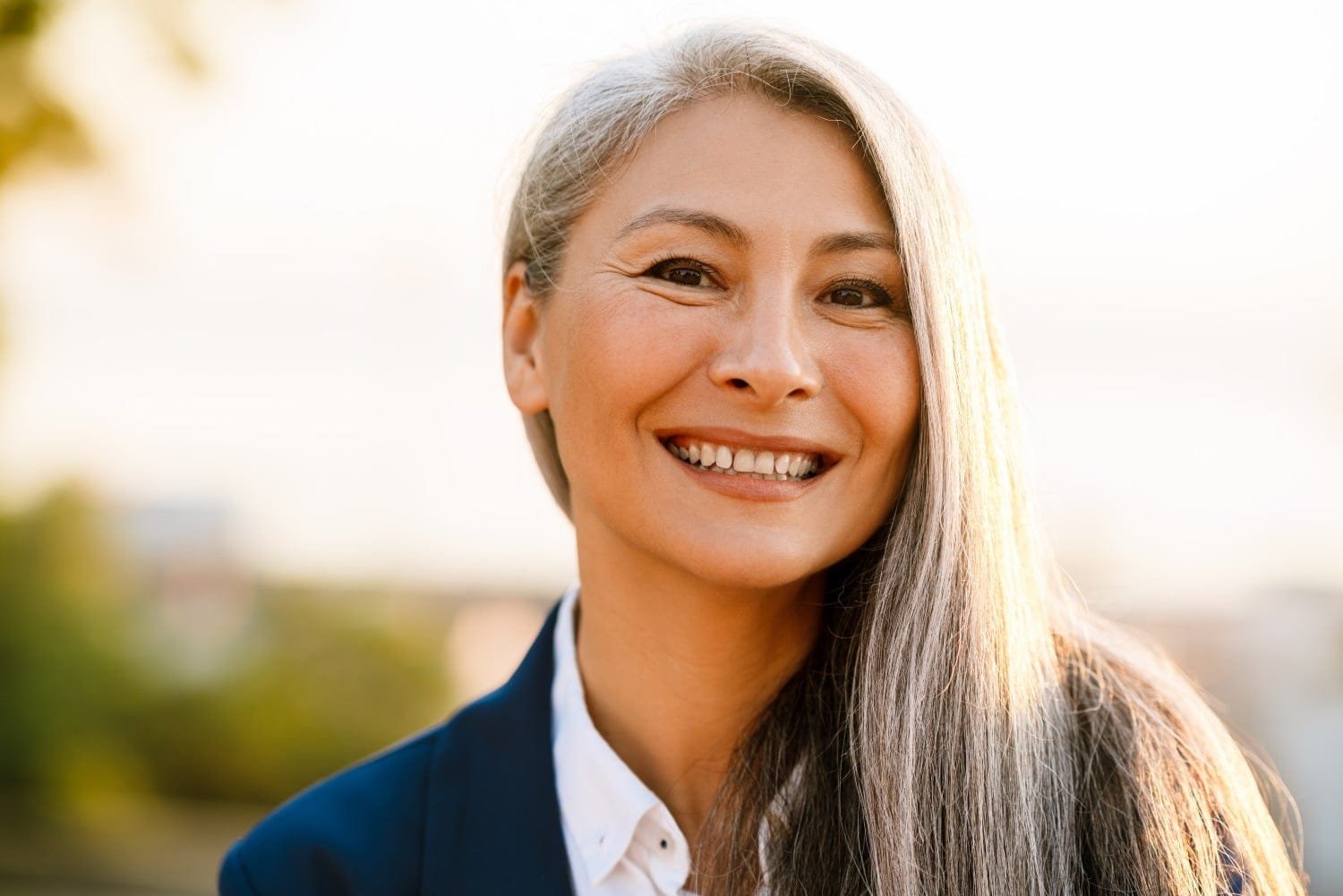 Frau mit langem grauem Haar, blauem Blazer und weißer Bluse bis zum Brustbereich fotografiert, lächelt zufrieden. Verschwommener Hintergrund mit Bäumen und trüben Himmel.