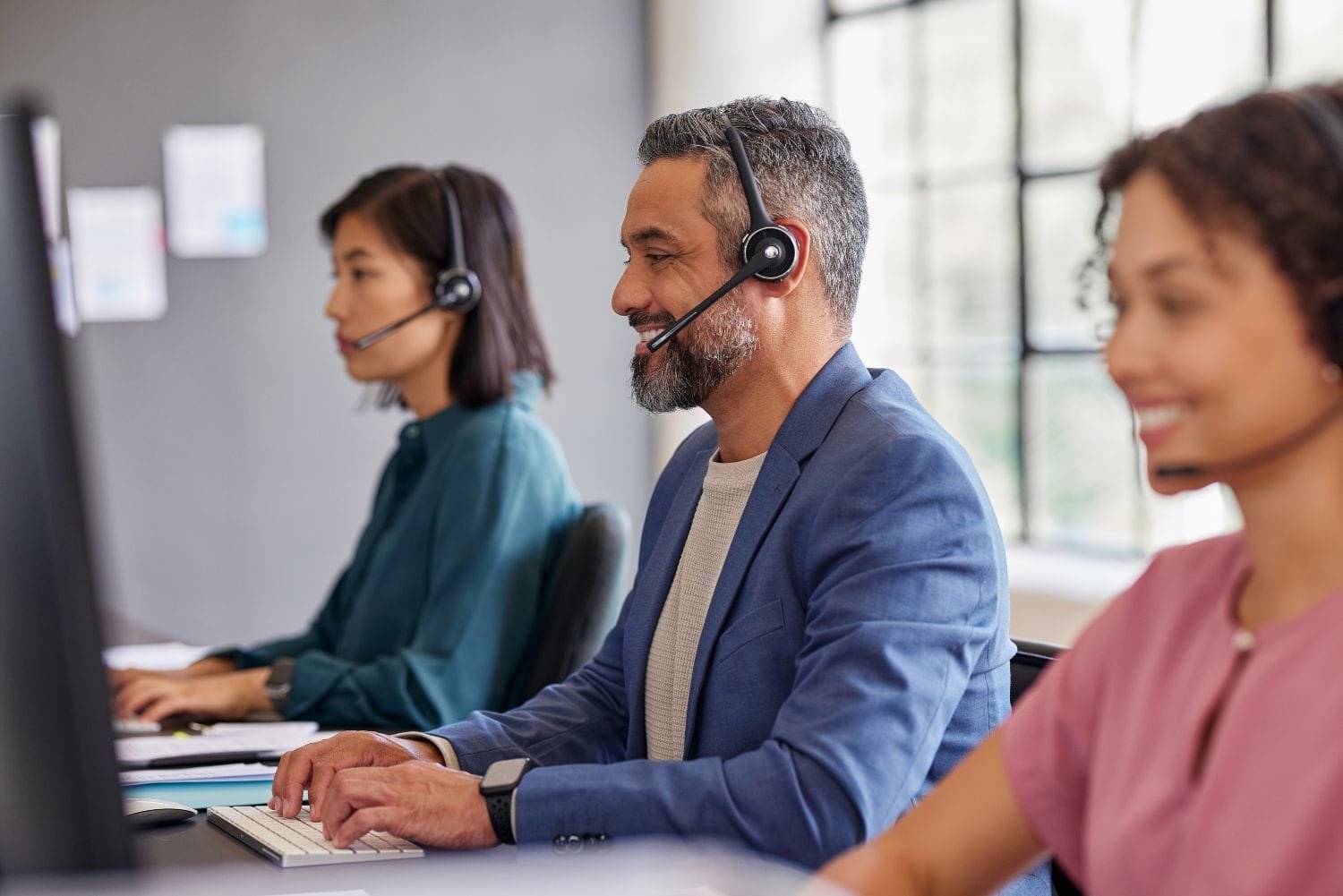 Drei Kundenservicemitarbeiter mit Headset auf dem Kopf in einer Reihe jeweils vor einem Bildschrim. Zwei Frauen und in der Mitte ein Mann, der lächelt.