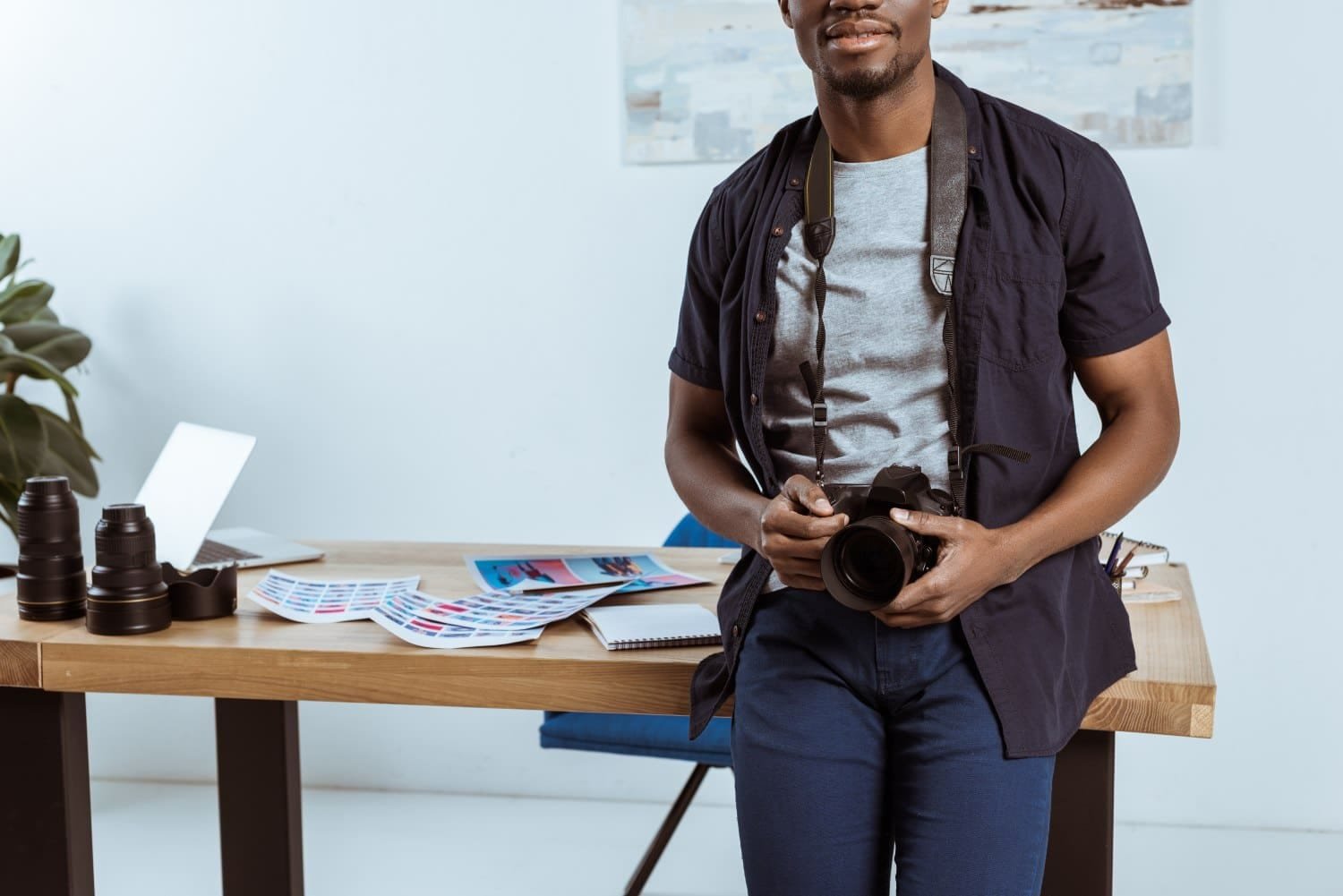 Mann in blau-grauer Kleidung und Fotoapparat um den Hals vom Bein bis zur Nase fotografiert, lehnt an Schreibtisch mit Dokumenten, Laptop und Kamerazubehör. Zimmerpflanze und weiße Wand im Hintergrund.