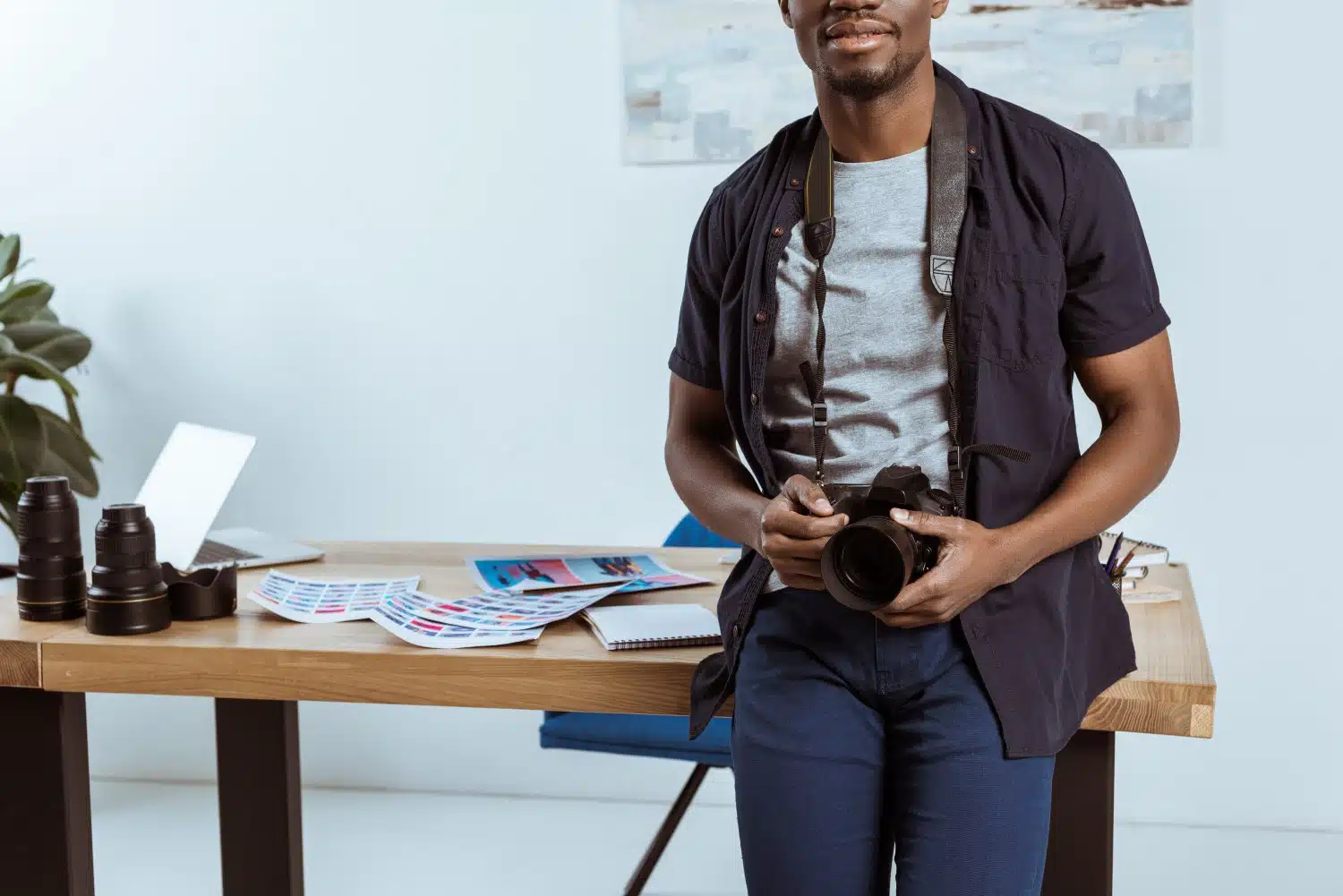 Mann in blau-grauer Kleidung und Fotoapparat um den Hals vom Bein bis zur Nase fotografiert, lehnt an Schreibtisch mit Dokumenten, Laptop und Kamerazubehör. Zimmerpflanze und weiße Wand im Hintergrund.