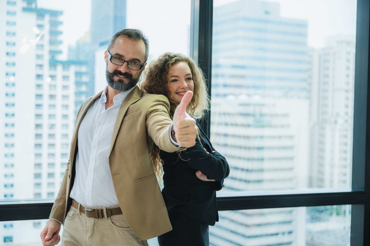 Ein lächelnder Mann mit Bart, Brille und im braunen Anzug hält den Daumen hoch. Rücken an Rücken steht eine lächelnde Frau in dunklem Anzug und mit gekreuzten Armen. Bodentiefe Fenster mit schwarzem Rahmen und Skyline-Ausblick im Hintergrund.