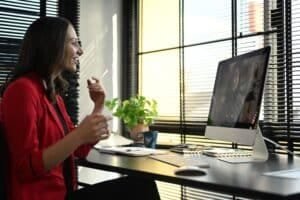 Lächelnde, brünette Frau in rotem Blazer und schwarzer Hose sowie Oberteil sitzt am Schreibtisch und führt einen Videocall mit einer Frau auf großem PC-Bildschirm. Fenster mit schwarzen Rollos um die erste Frau herum. Auf dunklem Schreibtisch ist außerdem eine Tastatur mit Maus, Topfpflanze, Kaffeetasse und Notizbuch samt Kugelschreiber.