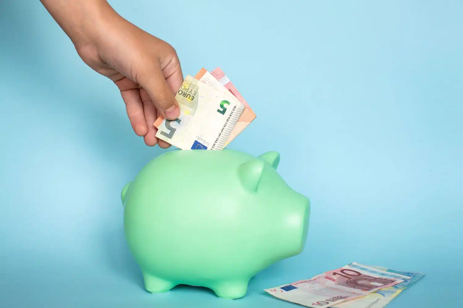 Hand, die Euro-Scheine in ein grünes Sparschwein legt. Daneben liegen ein 10- sowie ein 20-Euro-Schein leicht gefächert aufeinander. Blauer Hintergrund.