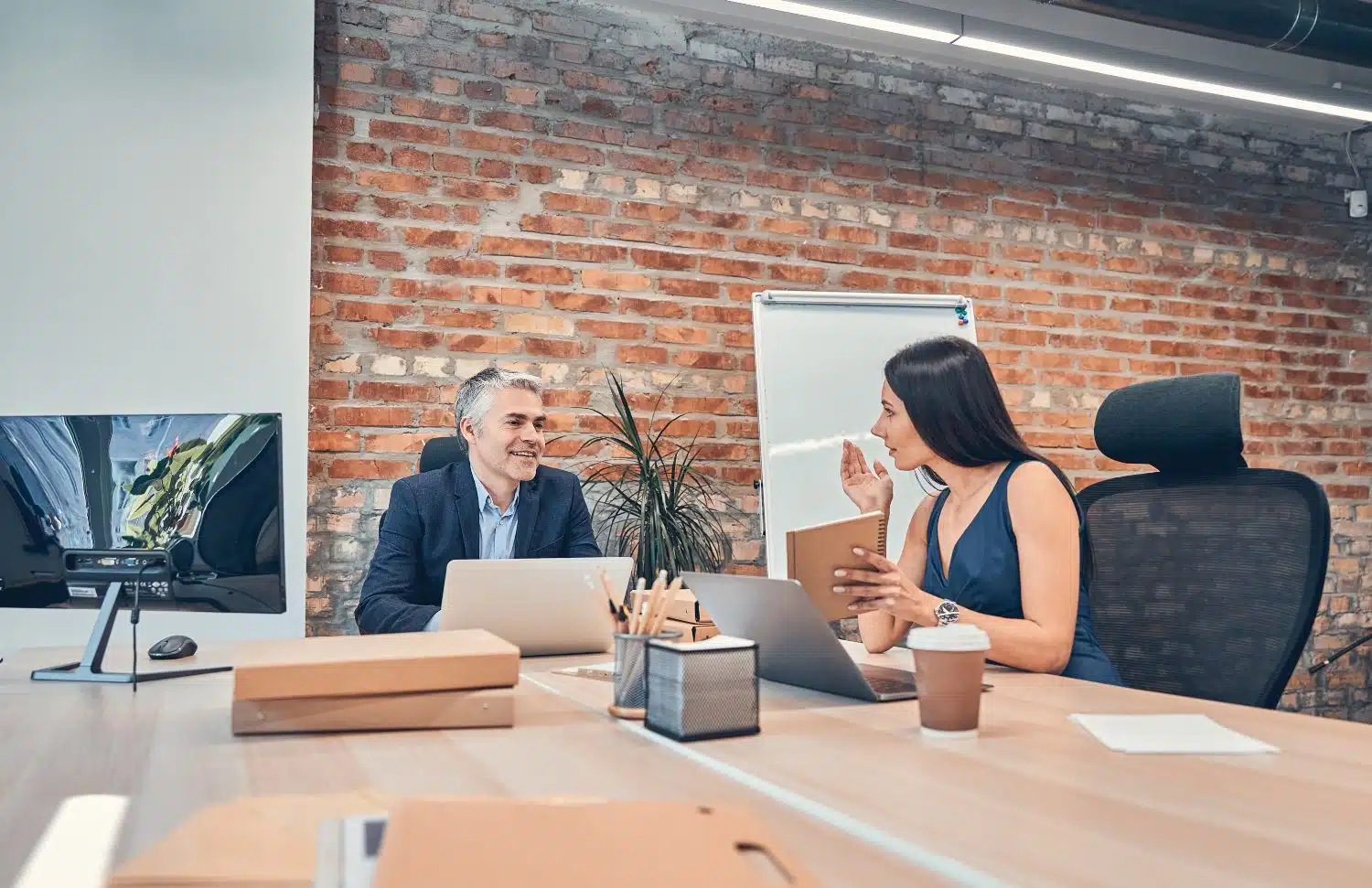Ein Mann und eine Frau in Business-Kleidung sitzen sich um die Ecke an Schreibtisch gegenüber. Büroutensilien, Bildschirm und Coffee-to-go auf Schreibtisch. Im Hintergrund Zimmerpflanzen und Ziegelsteinwand.