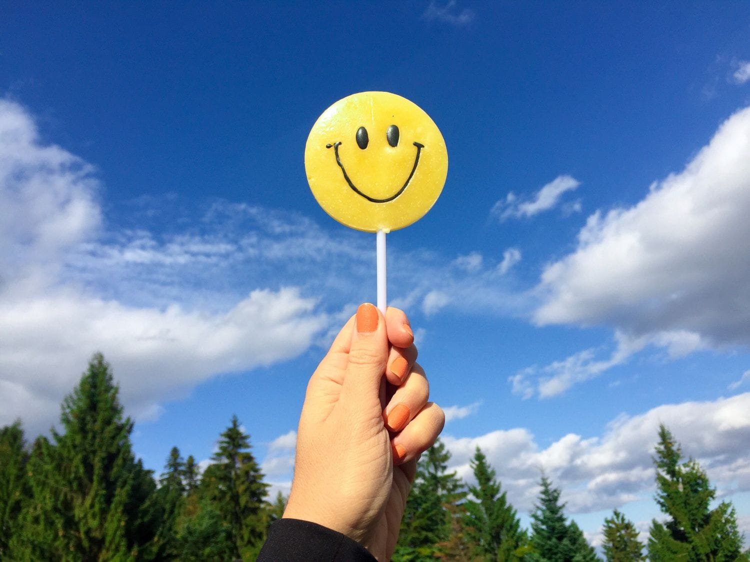 Frauenhand mit orange lackierten Nägeln und schwarzem Ärmel hält Lutscher mit gelbem Smiley-Gesicht hoch. Blauer Himmel mit Wolkenfetzen und Nadelbäumen im Hintergrund.