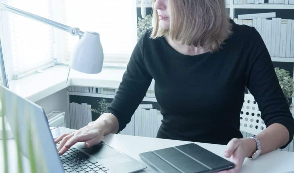 Eine Frau arbeitet an einem Laptop mit einem Tablet auf dem Schreibtisch in einem gut beleuchteten, modernen Arbeitsbereich.
