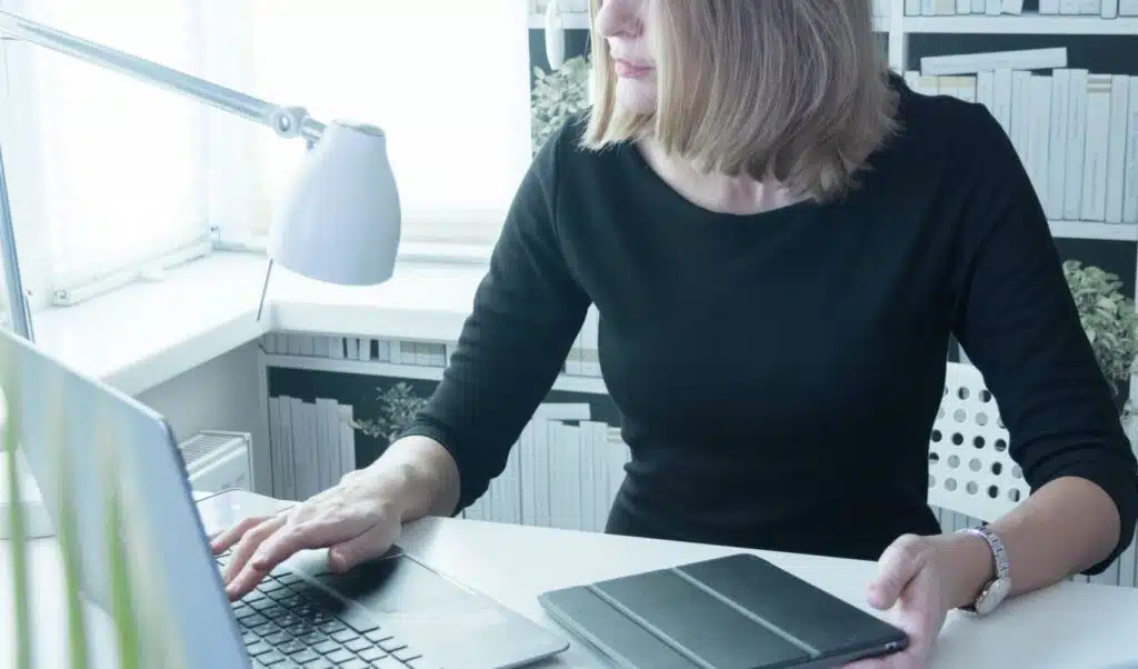 Eine Frau arbeitet an einem Laptop mit einem Tablet auf dem Schreibtisch in einem gut beleuchteten, modernen Arbeitsbereich.
