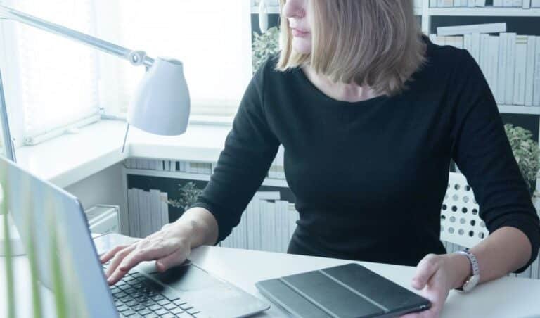 Eine Frau arbeitet an einem Laptop mit einem Tablet auf dem Schreibtisch in einem gut beleuchteten, modernen Arbeitsbereich.