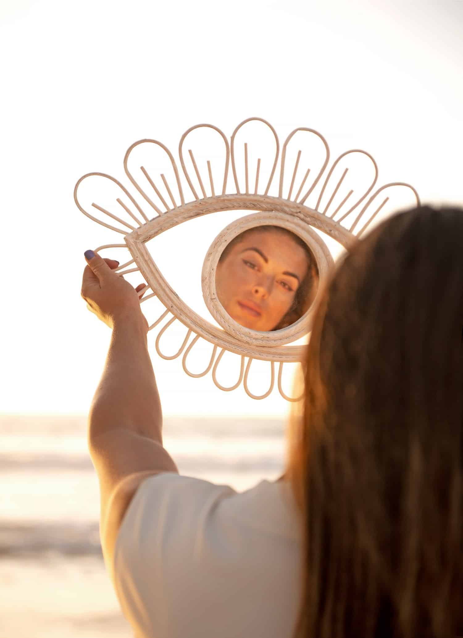 Frau von hinten mit hellbraunen Haaren und weißem T-Shirt am Strand hält einen augenförmigen Spiegel mit ausgestreckten Armen und betrachtet sich.