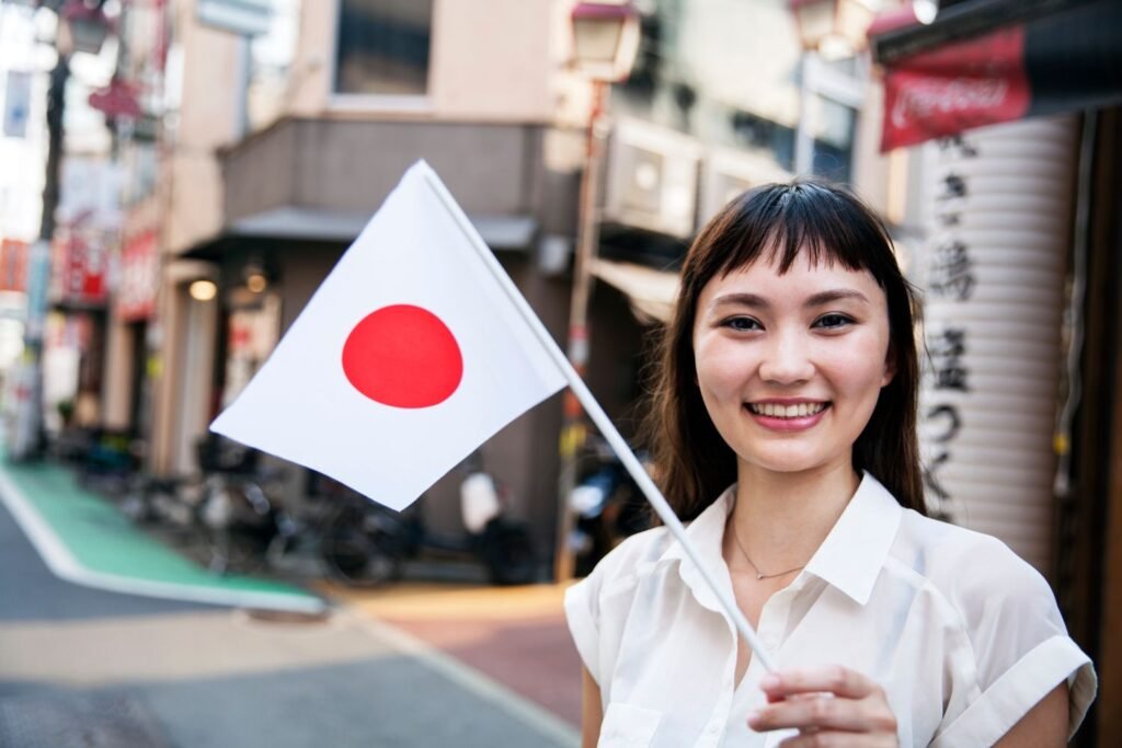 Lächelnde, brünette Frau in weißem T-Shirt hält kleine Japan-Flagge in der Hand, im Hintergrund Straße und mehrere Gebäude aneinandergereiht.