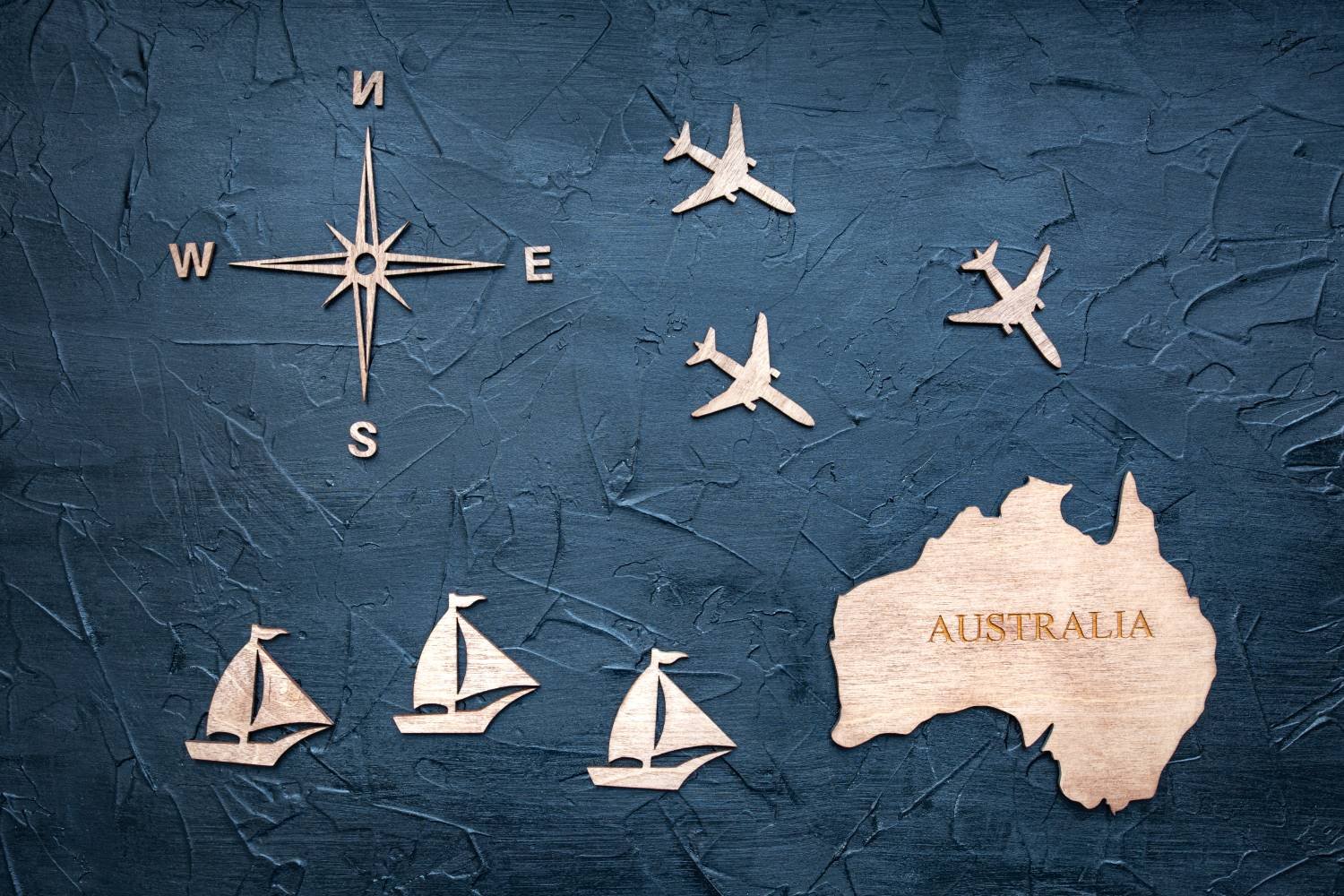 Blauer Hintergrund aus gewelltem Papier, darauf Holzausschnitte von Fluzeugen, Schiffen, einem Kompass und Australien.