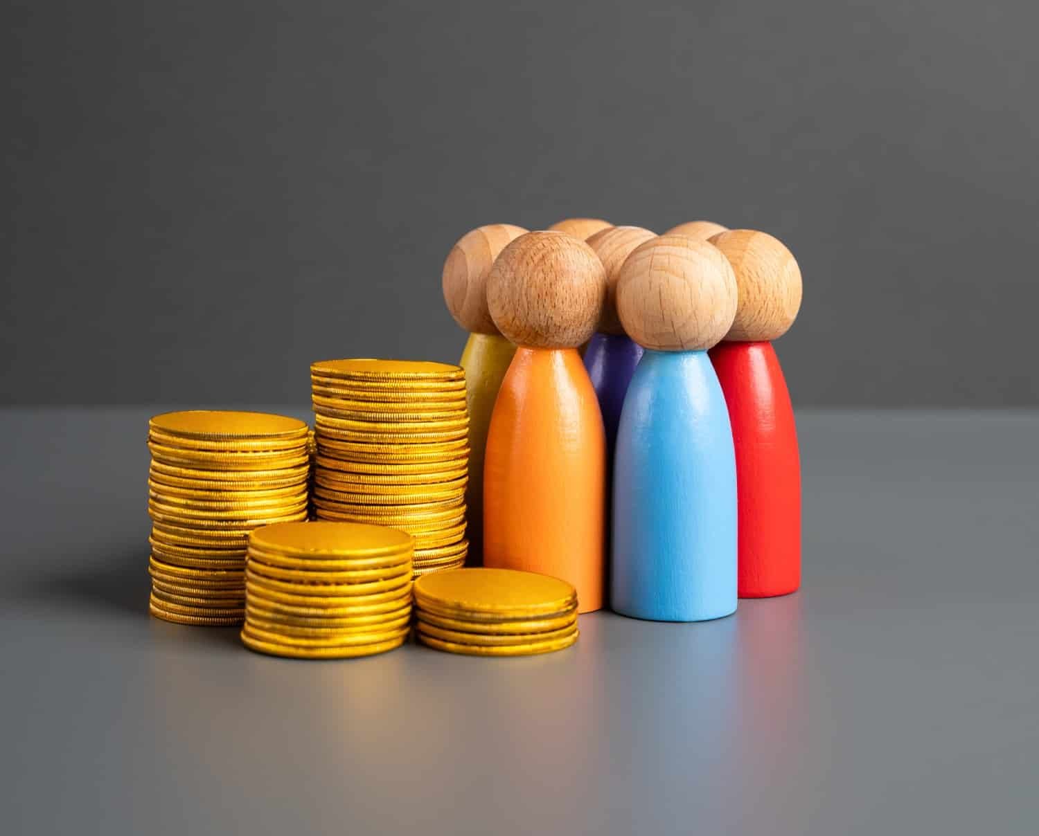 Grauer Hintergrund, ein Stapel goldener Münzen angelehnt an sieben bunten Brettspielfiguren aus Holz in einem Kreis zusammen.