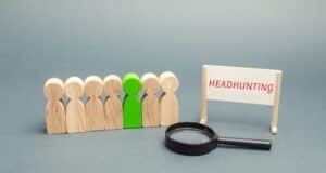 Sieben Holzfiguren aneinandergereiht auf grauem Grund, eins davon ist grün angemalt. Daneben ein kleiner Stand mit dem Schriftzug in Rot: Headhunting, und eine Lupe.