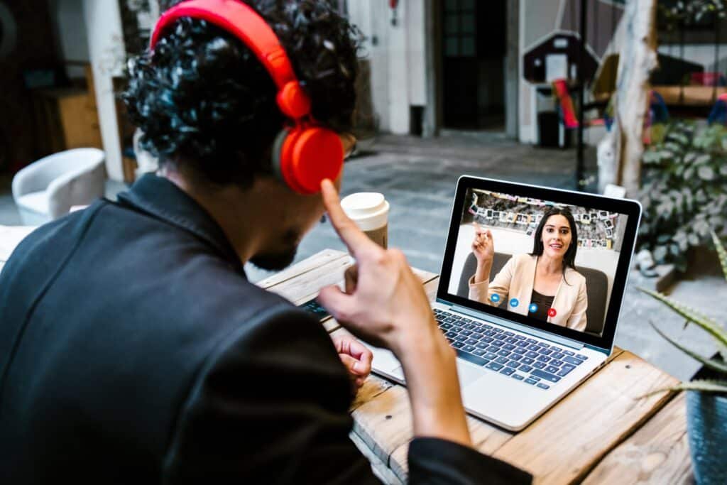 Ein Mann sitzend von hinten mit dunklem Haar und in dunklem Hemd am Schreibtisch und mit rotem Headset auf den Ohren. Auf Schreibtisch ein Laptop mit Frau in hellem Blaser und dunkler Bluse. Im Hintergrund Zimmer mit dunklem Boden und Einrichtung, darunter Zimmerpflanzen.