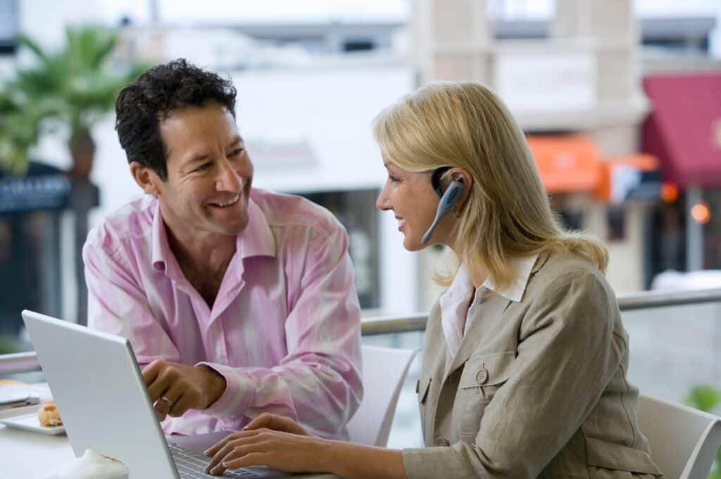 Dunkelhaariger Mann im rosa-weiß gestreiften Hemd und eine blonde Frau mit Headset auf Kopf und im beigefarbenen Hemd mit weißem Kragen an Schreibtisch vor Laptop. Stadt mit Palme im Hintergrund.