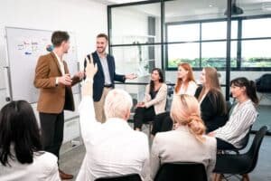 Eine Gruppe von Männern und Frauen in Businesskleidung – wovon einer die Hand hebt – sitzt auf schwarzen Stühlen im Halbkreise vor zwei Männern in Businesskleidung, die vor einem Whiteboard etwas erklären.