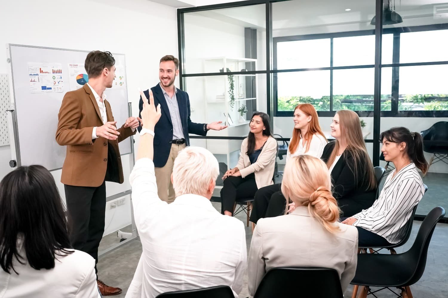 Eine Gruppe von Männern und Frauen in Businesskleidung – wovon einer die Hand hebt – sitzt auf schwarzen Stühlen im Halbkreise vor zwei Männern in Businesskleidung, die vor einem Whiteboard etwas erklären.