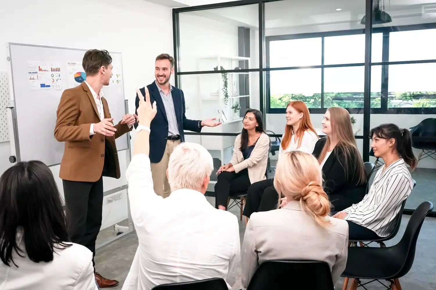 Eine Gruppe von Männern und Frauen in Businesskleidung – wovon einer die Hand hebt – sitzt auf schwarzen Stühlen im Halbkreise vor zwei Männern in Businesskleidung, die vor einem Whiteboard etwas erklären.