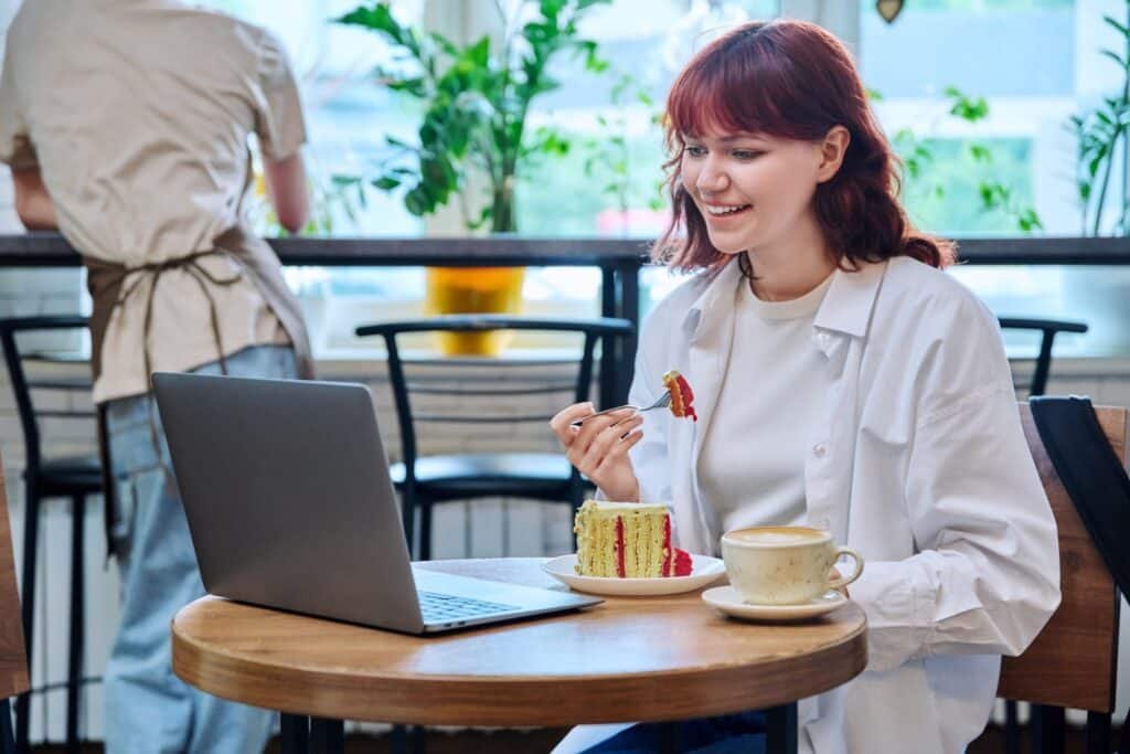 Lächelnde Frau mit dunkelrotem Haar, blauer Jeans und weißem, offenen Blazer sowie Top isst Kuchen an einem runden Tisch mit Laptop und Kaffeetasse drauf. Dahinter hohe Stühle und Person mit Schürze.