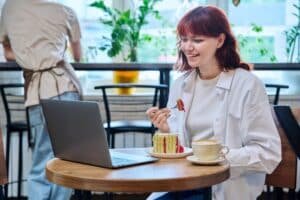 Lächelnde Frau mit dunkelrotem Haar, blauer Jeans und weißem, offenen Blazer sowie Top isst Kuchen an einem runden Tisch mit Laptop und Kaffeetasse drauf. Dahinter hohe Stühle und Person mit Schürze.