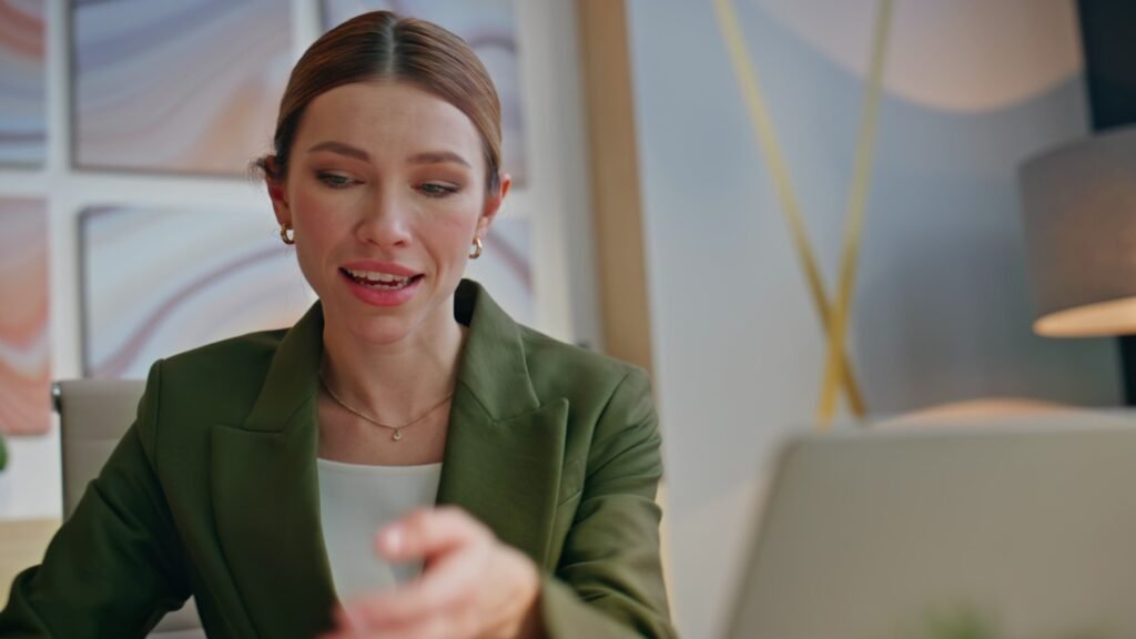 Frau mit dunkelblondem Zopf, grünem Blazer, weißer Bluse, goldenen Ohrringen und Kette spricht vor einem Laptop. Fenster und blaue Wand mit Goldakzenten im Hintergrund.