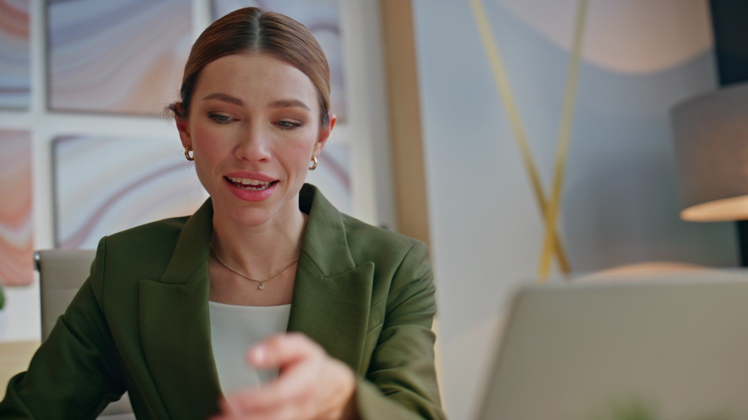 Frau mit dunkelblondem Zopf, grünem Blazer, weißer Bluse, goldenen Ohrringen und Kette spricht vor einem Laptop. Fenster und blaue Wand mit Goldakzenten im Hintergrund.