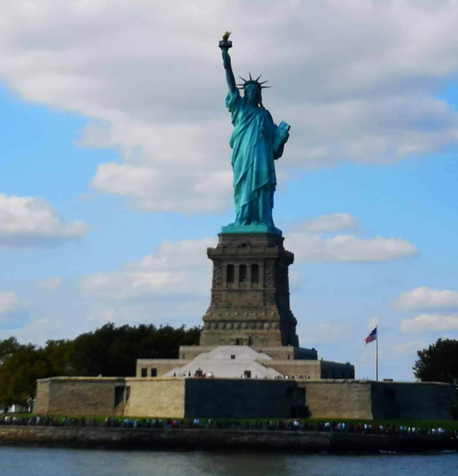 Freiheitsstatue vom Wasser aus fotografiert. Davor eine Menschenmasse, dahinter Flagge USA sowie Laubbäume. Darüber blauer Himmel mit Wolkenfetzen.